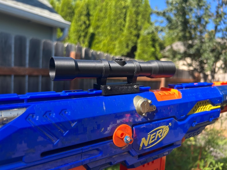 Puede incluir: Una mira telesc&oacute;pica de juguete cil&iacute;ndrica negra montada sobre un lanzador de juguete de pl&aacute;stico azul y naranja. El lanzador presenta el logotipo amarillo de NERF en el lateral, con un punto de fijaci&oacute;n circular naranja y un peque&ntilde;o detalle plateado.