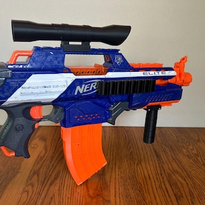 Puede incluir: Una pistola de juguete Nerf Rapidstrike CS-18 azul y naranja con una mira telescópica negra. La pistola de juguete tiene la palabra "NERF" impresa en el lateral. La pistola tiene un cargador naranja y detalles en naranja. La pistola está sobre una superficie de madera.