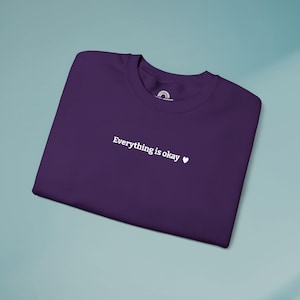 May include: A folded, dark purple t-shirt with the phrase "Everything is okay" printed in white, along with a small white heart symbol. The shirt is made of a soft material and is displayed on a light blue surface.