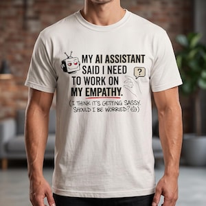 Puede incluir: Una camiseta blanca de manga corta con un gráfico de una cabeza de robot y el texto: MY AI ASSISTANT SAID I NEED TO WORK ON MY EMPATHY. (I THINK IT'S GETTING SASSY. SHOULD I BE WORRIED?) junto con un gesto de mano y un icono de signo de interrogación.