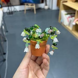 May include: A small orange plant pot filled with green moss and decorated with beaded flowers in green, white, and pink. The beaded flowers hang down the sides of the pot, giving a whimsical, handmade appearance.