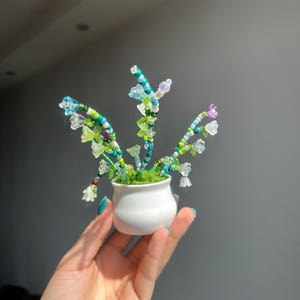 May include: A miniature potted plant featuring stems made of multi-coloured glass beads in shades of blue, green, white, and purple. The stems are topped with small, translucent flower-shaped beads and are set in a small, plain white ceramic pot.