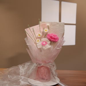 May include: A handcrafted bouquet of felt flowers in shades of pink and white, wrapped in delicate pink tissue paper and tied with a ribbon. The bouquet includes a pink rose, white and yellow daisies, and green stems.