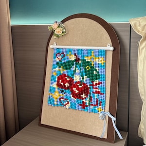May include: A beaded wall hanging featuring a cherry motif with red, green, and yellow beads on a light blue background. The piece is suspended from a white rod attached to an arched, brown-bordered display board adorned with a small floral accent.