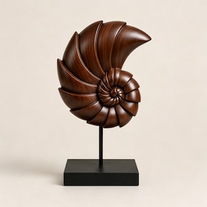 Puede incluir: Una escultura de madera de color marrón oscuro con forma de concha de nautilo y un diseño en espiral segmentado y liso. La concha está montada sobre una varilla metálica negra vertical y delgada, unida a una base rectangular negra sólida.