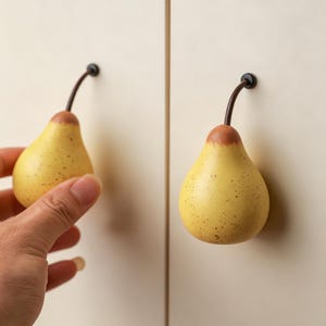 Peut inclure: Deux boutons de placard en forme de poire avec un corps jaune, des taches brunes et une tige brune. Les boutons sont fixés sur une surface claire, l'un d'eux étant tenu pour démontrer sa forme et sa taille en tant que quincaillerie fonctionnelle.