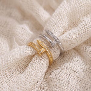 Può includere: Due anelli a forma di chiodo, uno color oro e uno color argento, ciascuno caratterizzato da una fila di piccole pietre chiare lungo la testa e parte del gambo. Gli anelli presentano un design avvolgente che imita un chiodo piegato.