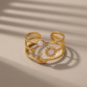 Può includere: Un anello aperto color oro con tre fasce parallele. Il design include un piccolo sole con un centro circolare rosa pallido, una luna crescente, una piccola stella e un accento circolare rosa pallido separato, integrati nella struttura.