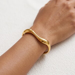 May include: A polished gold-tone bangle bracelet featuring a smooth, continuous wavy or undulating design. The metal surface is highly reflective, showcasing a curved, fluid shape that wraps around the wrist with a consistent, rounded profile.