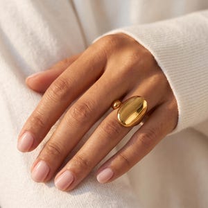 May include: A polished gold ring featuring a large, smooth, oval-shaped centrepiece and a smaller, rounded accent bead on the side. The ring has a high-shine, reflective metallic finish and a simple, continuous band design that sits comfortably on the finger.