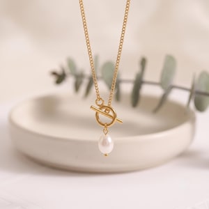 Op de afbeelding: Een goudkleurige ketting met een knevelsluiting aan de voorzijde. Een enkele, kleine, witte, ovaalvormige parelhangertje hangt aan het midden van de ronde knevelring, wat zorgt voor een eenvoudig en gebalanceerd sieraad.