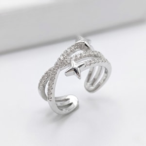 May include: A silver-toned open ring featuring a crossover band design. The ring is adorned with small, clear pavé-set stones along the bands and two polished, smooth silver-toned star accents positioned at the open ends of the ring structure.