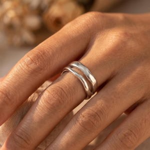 Puede incluir: Un anillo de tono plateado con un diseño de doble banda y un acabado liso y pulido. El anillo consta de dos bandas separadas, ligeramente onduladas e interconectadas que crean un espacio abierto entre ellas alrededor del dedo.