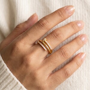May include: A gold-toned ring shaped like a bent nail, featuring a smooth polished band that wraps around the finger. The head and the pointed end of the nail design are encrusted with small, clear, sparkling stones along the top surface.