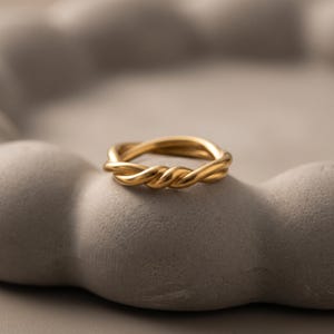 Könnte beinhalten: Ein goldfarbener Ring mit einem gedrehten, seilartigen Design. Das Band besteht aus mehreren ineinander verschlungenen Strängen, die eine durchgehende, strukturierte Schlaufe bilden, mit einer glatten, polierten Oberfläche, die das Licht reflektiert.