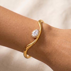 May include: A gold-toned bangle bracelet featuring a smooth, undulating wave-like band. The center of the bracelet is adorned with a single pear-shaped clear stone, surrounded by a delicate halo of tiny, sparkling white accent stones.
