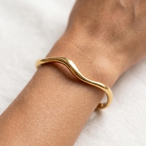 May include: A polished gold-tone cuff bracelet featuring a smooth, continuous wavy or serpentine design. The metal band has a rounded profile and a reflective surface that curves gently around the wrist, creating a fluid and minimalist aesthetic.