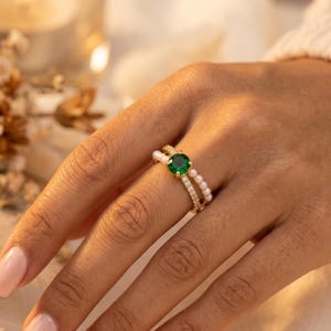 May include: A gold ring featuring a round, vibrant green center stone set in a prong setting. The band is split into two distinct rows: one row is adorned with small, white, spherical pearls, while the other row is encrusted with tiny, clear, sparkling stones.