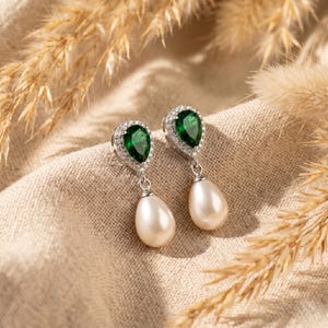 May include: A pair of elegant drop earrings. Each earring features a pear-shaped, emerald-green gemstone surrounded by small, clear crystals. Below the green stone hangs a large, white, teardrop-shaped pearl. The earrings are set on a silver-colored metal.