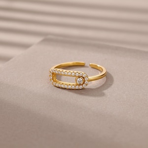 Puede incluir: Un anillo abierto de tono dorado con una pieza central ovalada alargada. El marco ovalado está incrustado con pequeñas piedras claras y brillantes, y una piedra redonda más grande y clara se sitúa en un extremo de la abertura ovalada.