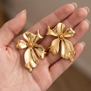 Puede incluir: Un par de pendientes dorados con forma de flor. Cada pendiente tiene cinco pétalos y un centro texturizado. Los pendientes se sostienen en una mano, mostrando su acabado metálico brillante. El diseño recuerda a un arreglo floral estilizado.