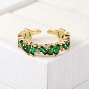 Può includere: Un anello aperto color oro caratterizzato da una serie di pietre rettangolari verde smeraldo taglio baguette alternate a piccole pietre rotonde trasparenti lungo la fascia. L'anello presenta un design aperto sul retro per la regolazione.