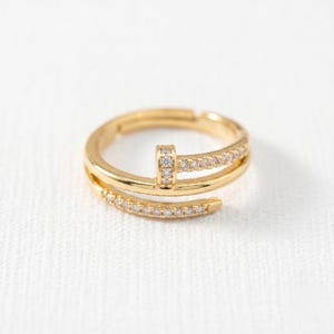 May include: A gold-toned open ring featuring a nail-inspired design with a flat head and a tapered end. The band is accented with small, clear, sparkling stones embedded along the surface, creating a continuous shimmering effect across the metal.