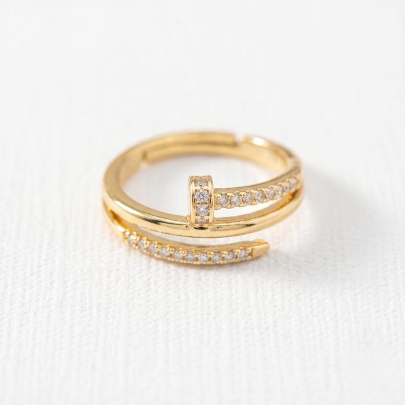May include: A gold-toned open ring featuring a nail-inspired design with a flat head and a tapered end. The band is accented with small, clear, sparkling stones embedded along the surface, creating a continuous shimmering effect across the metal.