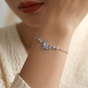 May include: A silver bracelet with a crescent moon pendant, finished in light blue enamel, and small star details. The bracelet has a delicate chain design, with additional star-shaped charms.
