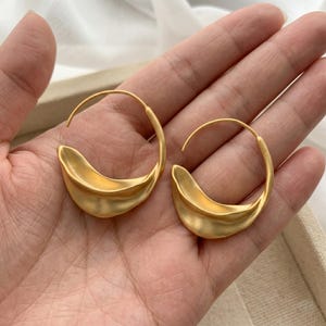 May include: A pair of gold-coloured hoop earrings. Each earring has a curved, crescent-shaped design with a matte finish. The earrings are held in a person's hand, showing their size and shape. The overall style is modern and minimalist.