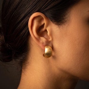 May include: A thick, semi-circular gold hoop earring with a brushed, matte finish. The earring features a wide, curved design that sits snugly against the earlobe, showcasing a smooth, metallic texture and a warm, golden tone throughout the piece.