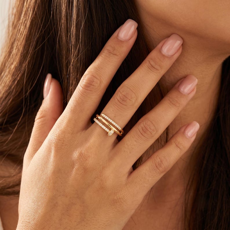 May include: A gold-toned ring shaped like a bent nail, featuring a double-band design. The surface of the ring is encrusted with small, clear, sparkling stones along the entire length of both bands, creating a continuous shimmering effect.