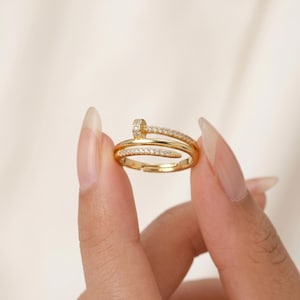 May include: A gold-toned ring shaped like a nail that wraps around the finger. The head of the nail and the band are encrusted with small, clear, sparkling stones. The ring features an open design, allowing for adjustment to fit different finger sizes.