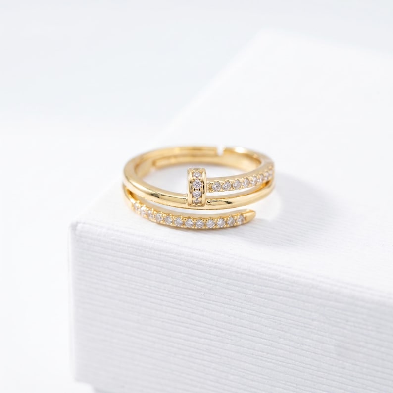 May include: A gold-toned ring featuring a bypass design with two overlapping bands. The ring is embellished with small, clear, sparkling stones set along the surface of both bands and a central vertical bar connecting the two ends.