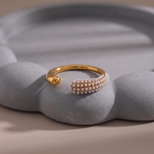 Könnte beinhalten: Ein goldfarbener offener Ring mit einer glatten, polierten Schiene auf der einen Seite und einem texturierten Bereich, der dicht mit kleinen, weißen, kugelförmigen Perlen besetzt ist, auf der anderen Seite. Das Design ist kreisförmig mit einer Lücke und zeigt einen Kontrast zwischen der metallischen Oberfläche und dem perlenbesetzten Detail.