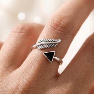 May include: An adjustable silver-toned ring featuring a textured feather design on one end and a black triangular inlay on the other end. The open-ended band is crafted with a polished finish, showcasing a contrast between the metallic silver and the dark black geometric shape.