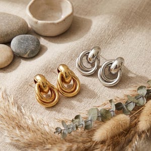 May include: Two pairs of earrings, one gold-toned and one silver-toned, are displayed on a neutral linen surface. The earrings feature a linked design with a rounded top and a looped bottom. Decorative elements include dried grasses, stones, and a small ceramic bowl.
