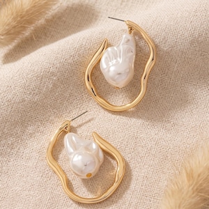 May include: A pair of gold-toned hoop earrings with an irregular, wavy frame. Each earring features a single, lustrous white baroque-style pearl suspended within the centre of the open, organic-shaped metal loop, secured with a standard post back.