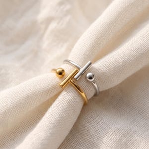 May include: A two-tone open ring featuring a gold-coloured band with a horizontal bar and a small sphere, layered with a silver-coloured band that also includes a horizontal bar and a small sphere. The metal surfaces are smooth and polished.