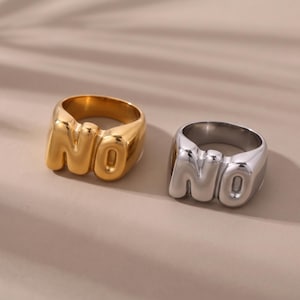 May include: Two chunky rings, one in a polished gold tone and one in a polished silver tone, each featuring the letters NO in a rounded, three-dimensional bubble font style integrated into the band design with a smooth, reflective metallic finish.