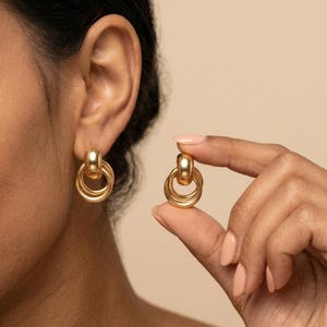 Peut inclure: Boucles d'oreilles dorées avec une tige et trois anneaux connectés. Une boucle d'oreille est portée à l'oreille, l'autre est tenue par une main. Les boucles d'oreilles ont une finition métallique brillante et un design classique.