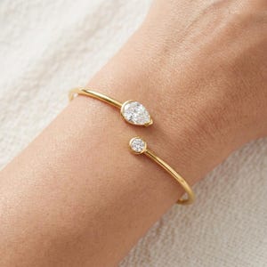 Op de afbeelding: Een open gouden armband met twee uiteinden, waarvan de ene is versierd met een peervormige, heldere edelsteen en de andere met een kleinere, ronde, heldere edelsteen. De dunne, gepolijste gouden band zorgt voor een verfijnde uitstraling.