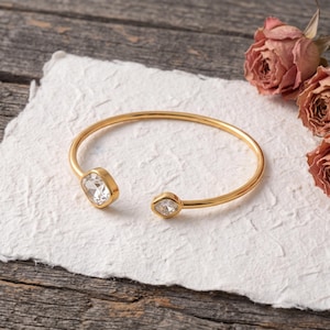 May include: A gold-toned open cuff bracelet featuring two clear, faceted, square-shaped crystals at each terminal end. The bracelet has a smooth, polished metal band that curves into a circular shape, highlighting the reflective quality of the stones.