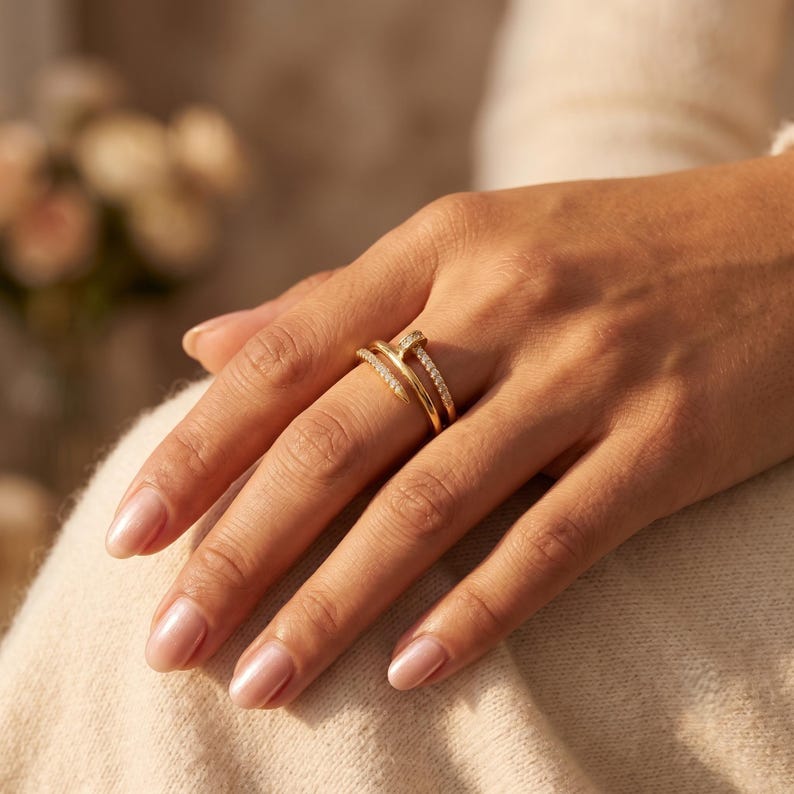 May include: A gold-toned ring featuring a nail-inspired design that wraps around the finger. The band is split into two parallel sections, with one section encrusted with small, clear, sparkling stones along its entire length.