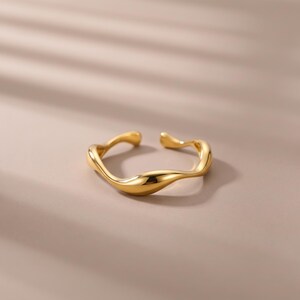 Peut inclure: Une bague ouverte dorée présentant un design de bande lisse, ondulé et semblable à une vague. La surface métallique est polie et réfléchissante, créant une forme continue et fluide qui reste ouverte sur le dessus pour un ajustement.