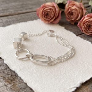 Puede incluir: Una pulsera plateada que presenta dos pequeños cubos metálicos, un anillo circular y un eslabón ovalado conectados a una fina cadena de cuentas de múltiples hilos. La pieza de joyería está hecha de metal plateado pulido brillante.