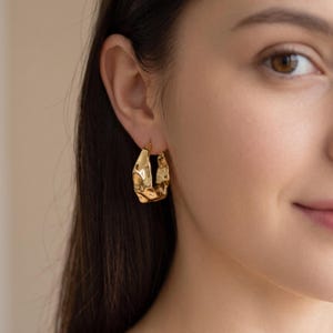 May include: A gold hoop earring with a textured, irregular, and organic surface. The hoop has a polished, reflective finish that catches the light, creating highlights and shadows across its uneven, molten-like metal form on an earlobe.