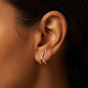 May include: A gold ear cuff featuring an X-shaped design. One side of the cross is smooth polished gold, while the other side is encrusted with a row of small, clear, sparkling stones. The piece wraps around the outer edge of the ear cartilage.