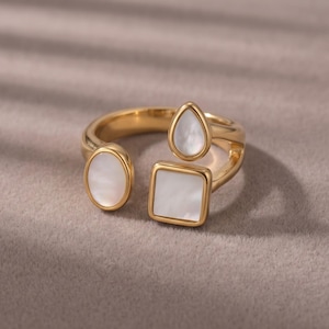 Puede incluir: Un anillo abierto de tono dorado con tres engastes geométricos distintos, cada uno incrustado con un acento de nácar blanco liso e iridiscente. El anillo incluye una incrustación ovalada, una cuadrada y una en forma de lágrima en una banda pulida.