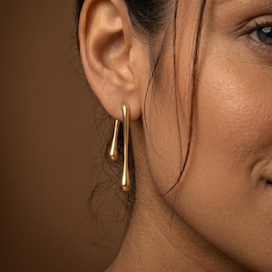 May include: Gold-colored, teardrop-shaped earrings. The earrings have a long, slender post with a rounded teardrop shape at the bottom. The earrings are worn on a person's ear, with the post extending downward. The earrings are made of a shiny, reflective metal.